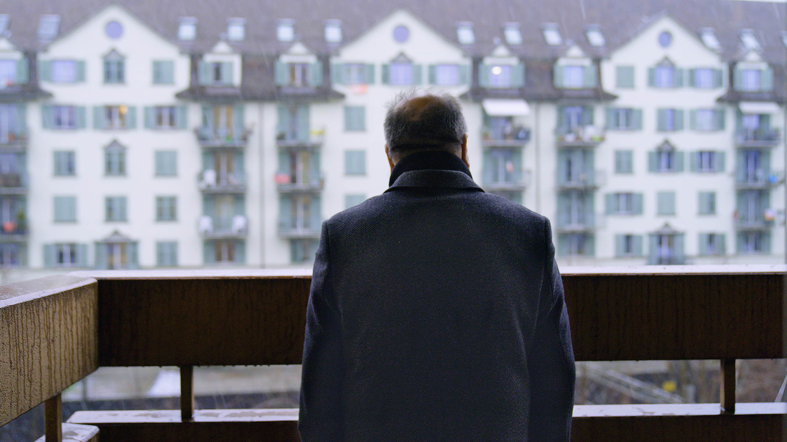 Un homme debout sur son balcon regarde une rangée d’immeubles en face de lui.