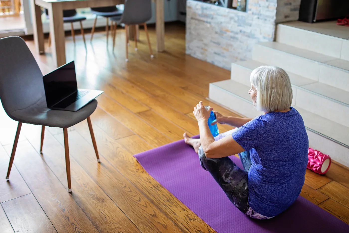 Bild eine Frau sitzt im Wohnzimmer auf einer Yogamatte und öffnet eine Wasserflasche. Daneben steht auf einem Stuhl ein aufgeklappter Laptop.