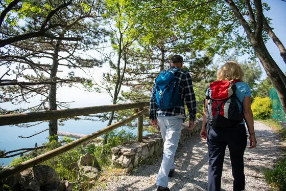 Bild Ein Paar mit Rucksäcken wandert  über einen flachen schattigen Weg hoch über einem See.