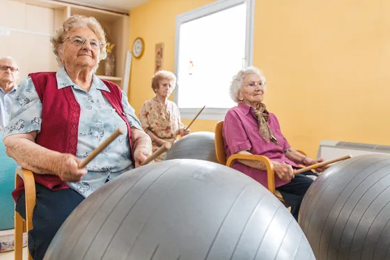 Bild alte Personen sitzen auf einem Stuhl und haben einen silberfarbigen Gymnastikball vor sich. In den Händen halten sie Trommelschläger und trommeln im Rhytmus auf den Ball.