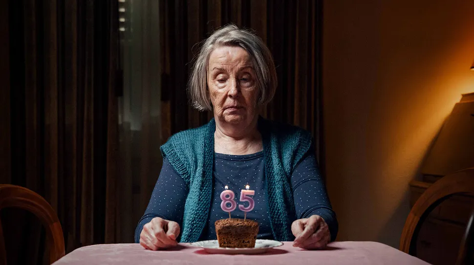 Eine alte Frau sitzt am Esstisch, vor sich hat sie ein kleines Kuchenstück, sie muss ihren 85. Geburtstag leider alleine verbringen.