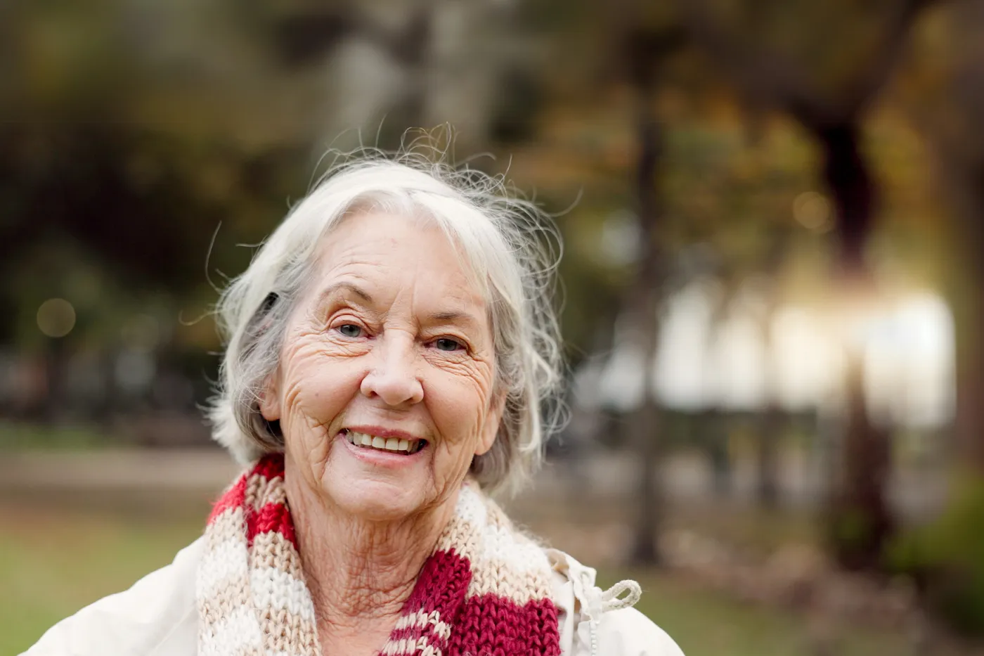 Das Foto zeigt eine Frau mit kurzen weissen Haaren, die in die Kamera lächelt. Sie trägt einen weiss-rot gemusterten Wollschal, während der Hintergrund unscharf ist.