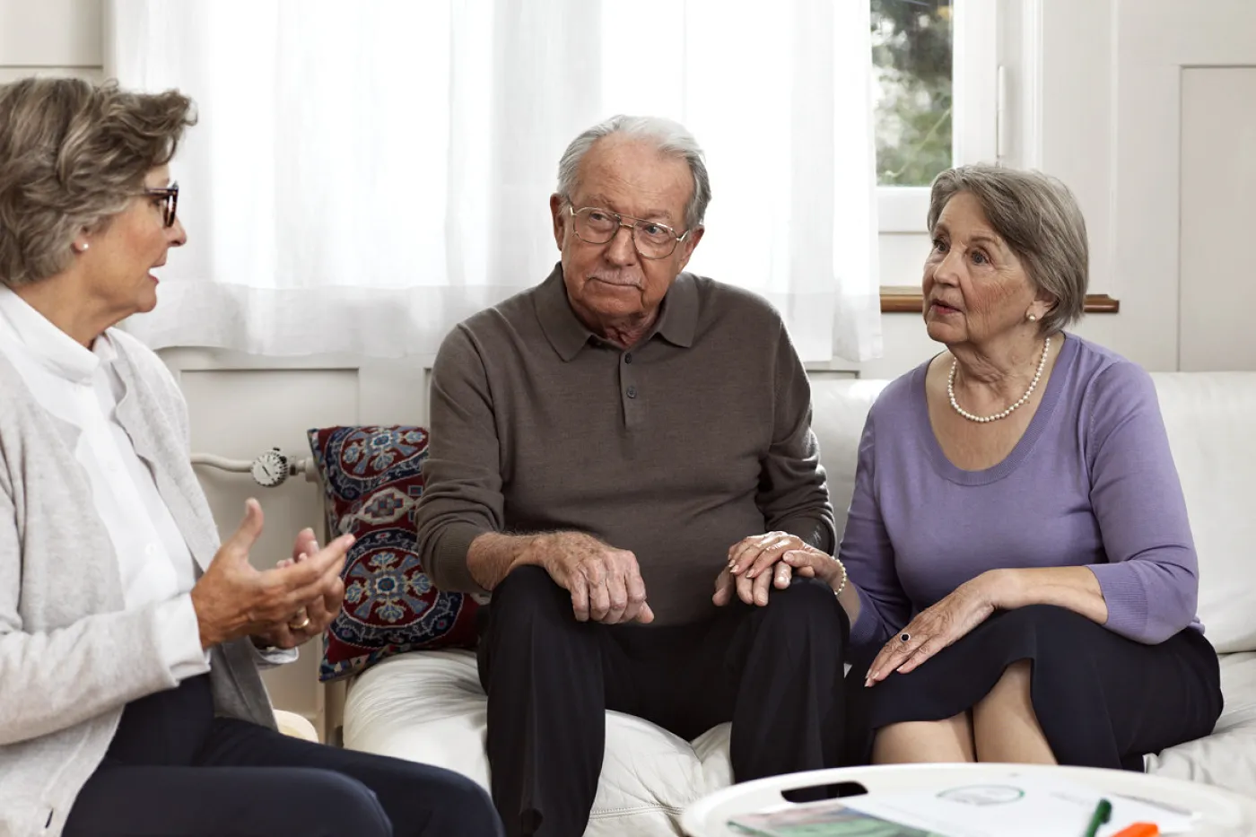 Bild ein Ehepaar sitzt auf dem Sofa. Die Frau hat die Hand auf die des Mannes gelegt. Sie hören einer Frau zu, die ihnen etwas erklärt.