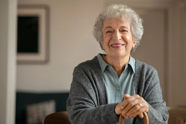 Bild Eine Seniorin sitzt auf einem Stuhl, streckt die Arme aus und hält mit beiden Händen einen Stock fest.