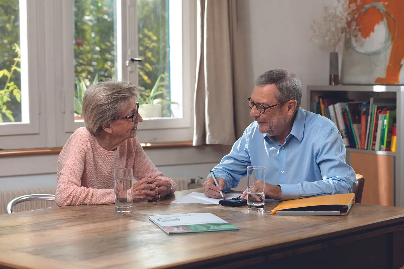 Bild eine Frau und ein Mann sitzen an einem Holztisch auf dem ein Ordner und zwei Gläser stehen. Sie sprechen zusammen und der Mann füllt ein Formular aus.