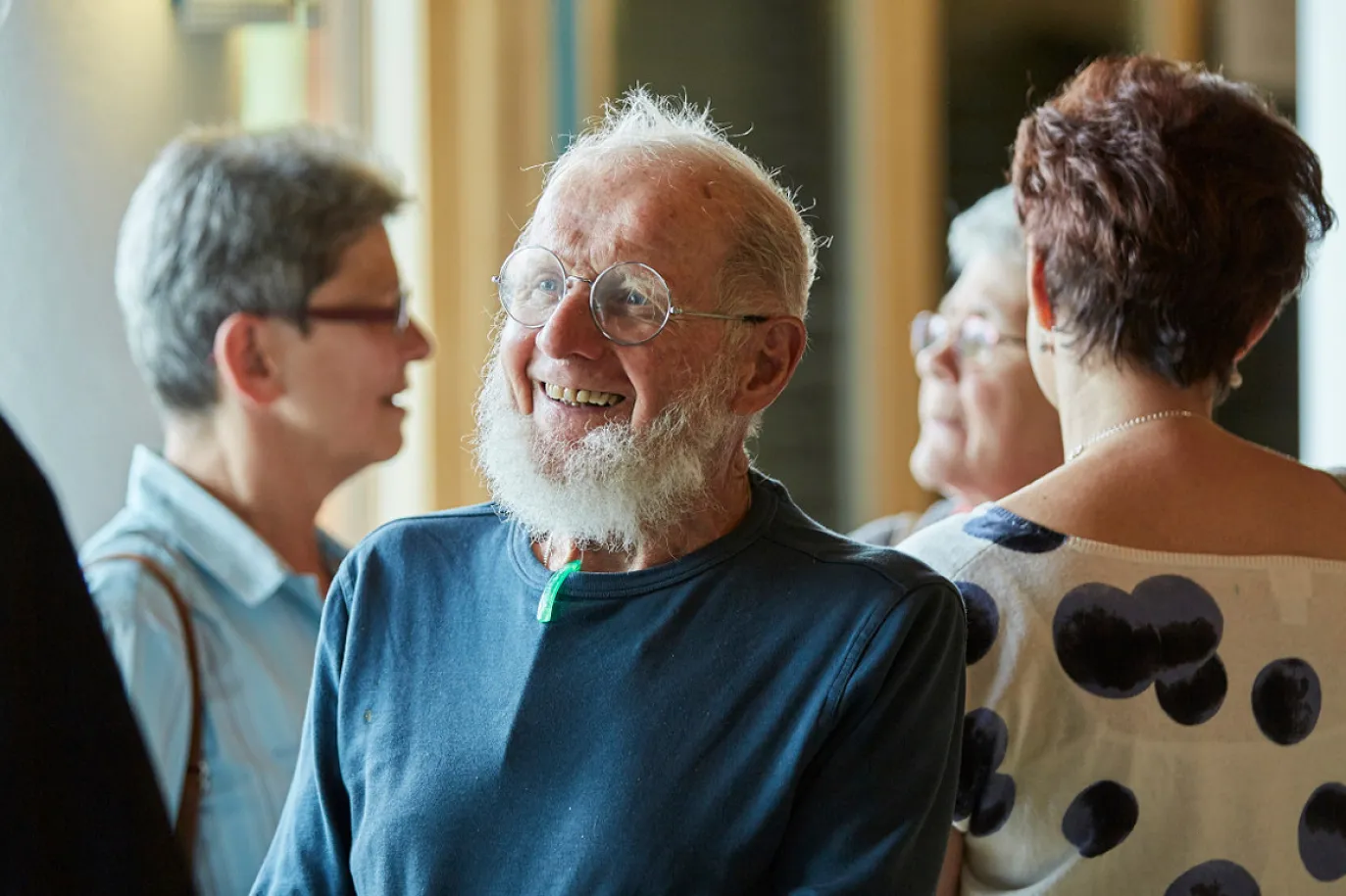 Image au premier plan d'un homme âgé souriant avec des lunettes rondes et une barbe blanche. A l'arrière-plan, trois femmes discutent.