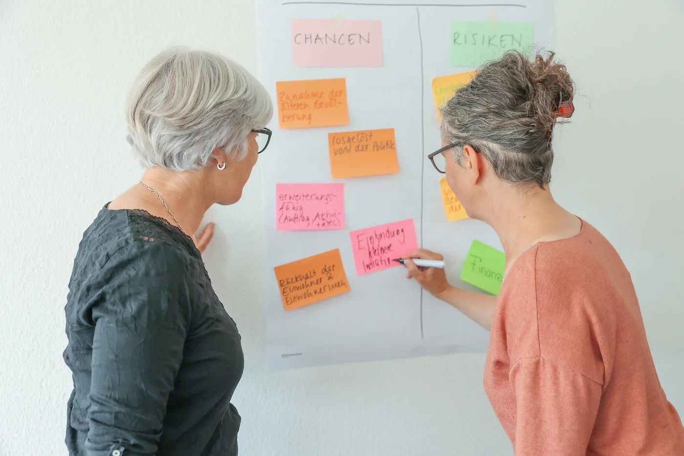 Deux femmes discutent des notes de différentes couleurs sur un tableau à feuilles mobiles.
