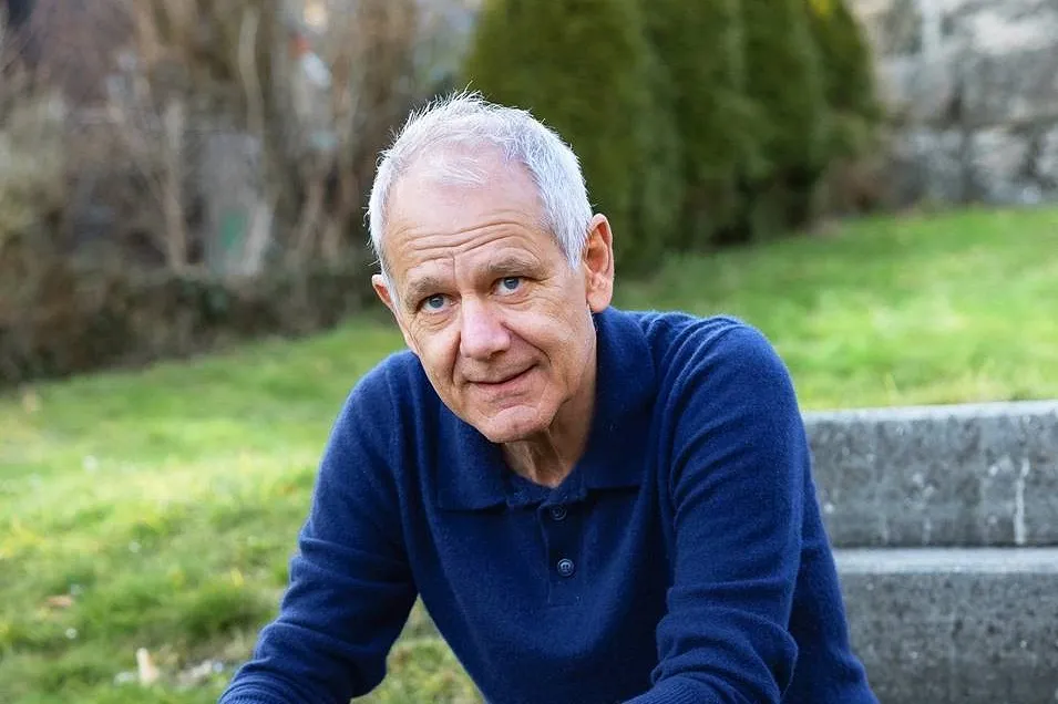 Un homme âgé est assis dehors sur un escalier en pierre, un smartphone à la main, et regarde l'appareil photo avec un air sympathique. En arrière-plan, on aperçoit une pelouse, des buissons et une clôture.