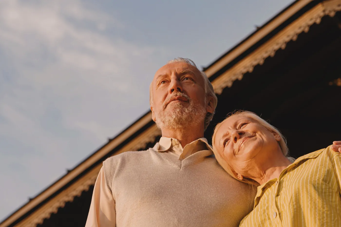 Image un homme a passé son bras autour de l'épaule d'une femme. Elle appuie sa tête contre son épaule. Ils se tiennent devant une maison et ont l'air satisfaits.