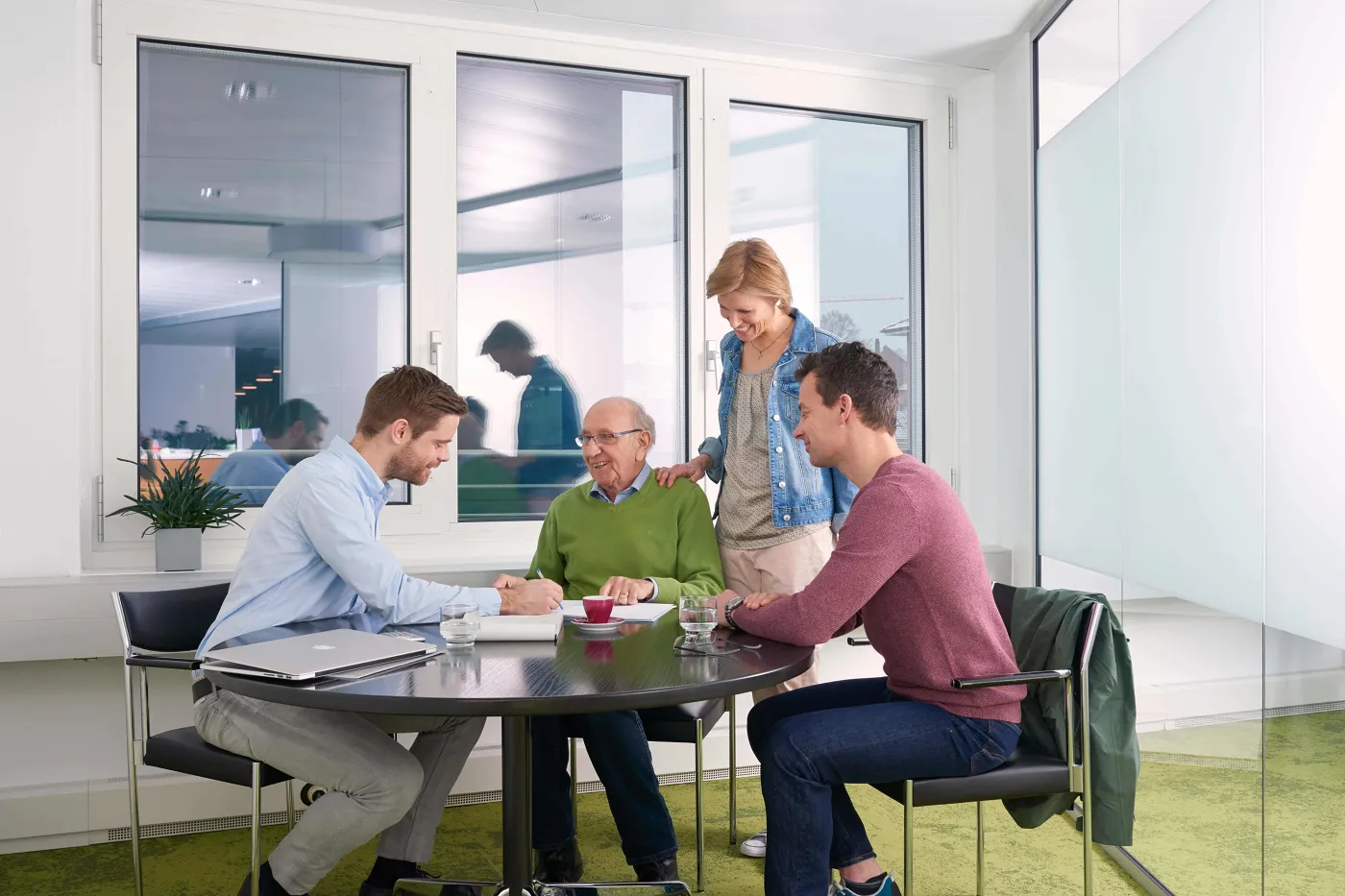 Image un homme âgé est assis avec deux jeunes hommes à une table ronde noire une jeune femme se tient à côté et a posé sa main sur l'épaule de l'homme âgé.