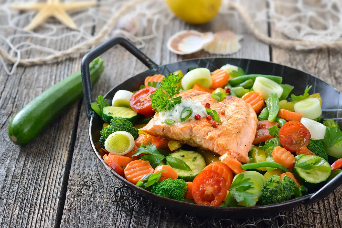 Photo une poêle avec différents légumes frais et un morceau de poisson posé dessus sur une table en bois. A côté, une courgette adore.