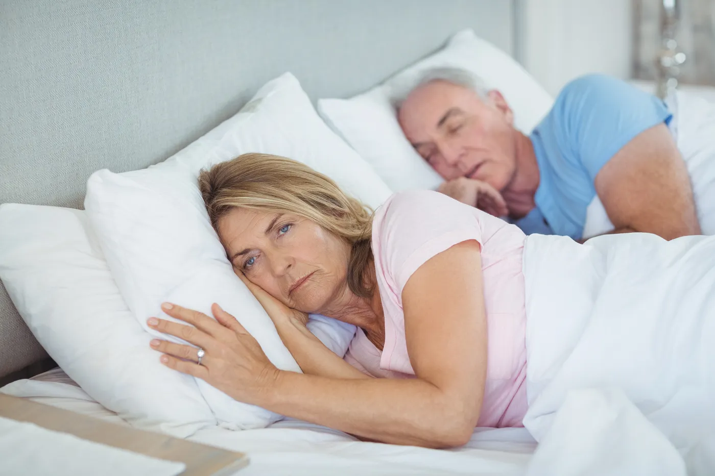 Photo d'un couple allongé sur le lit. Tous deux en position latérale. L'homme au fond dort, la femme au premier plan est allongée, éveillée, les yeux ouverts.