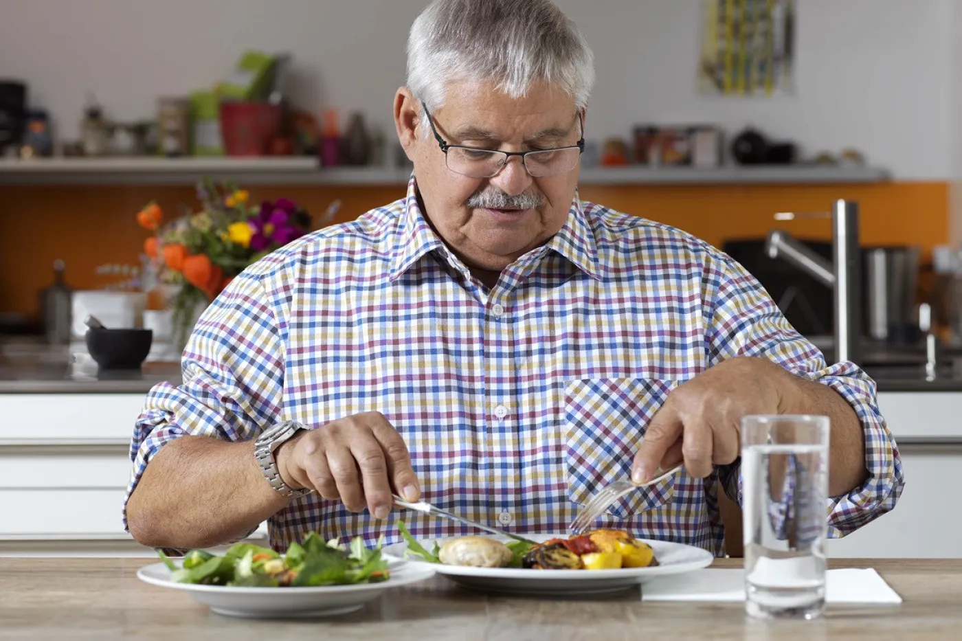 Image Un homme en chemise à carreaux est assis à table et mange de la viande et des légumes dans une assiette blanche. A côté, il y a une assiette de salade et un verre d'eau.