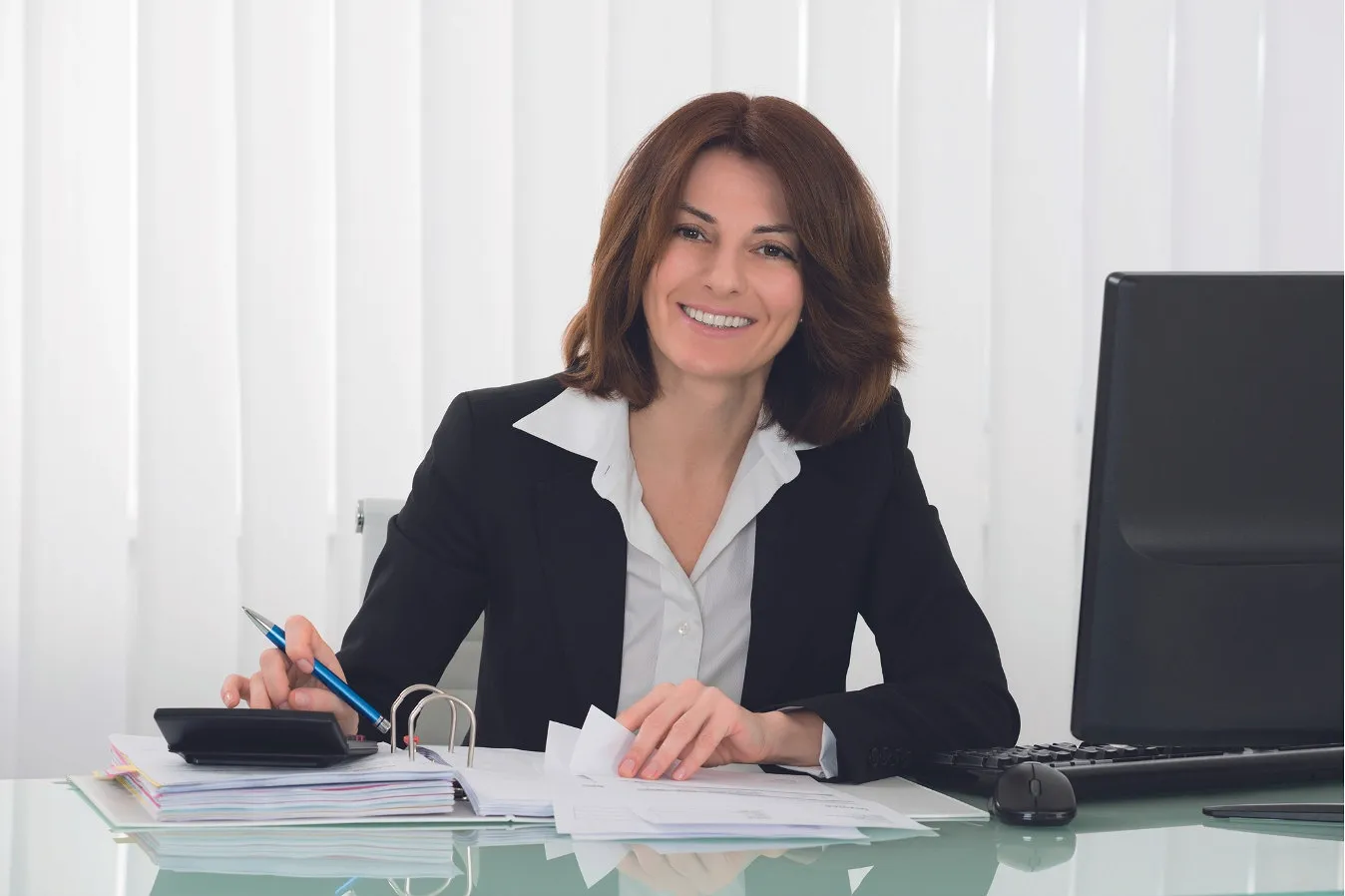 L'image montre une femme souriante avec des cheveux bruns jusqu'aux épaules, assise dans un bureau moderne. Elle porte un blazer noir et un chemisier blanc et semble professionnelle.