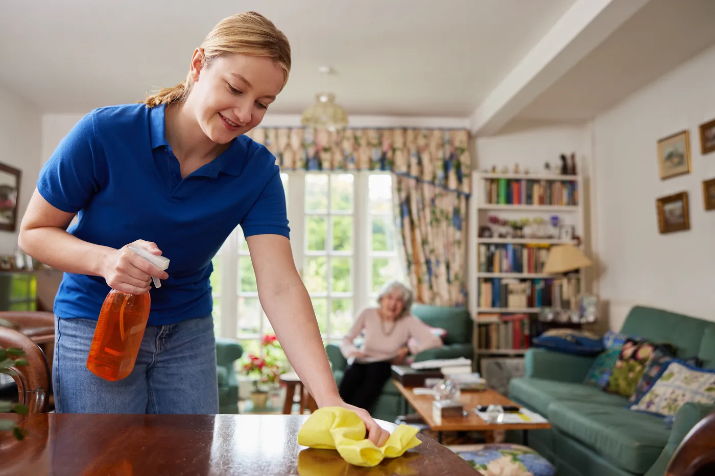 Image une jeune femme nettoie une table en bois avec un chiffon jaune. Dans l'autre main, elle tient un vaporisateur. En arrière-plan, une femme plus âgée, assise dans un fauteuil vert, la regarde faire.